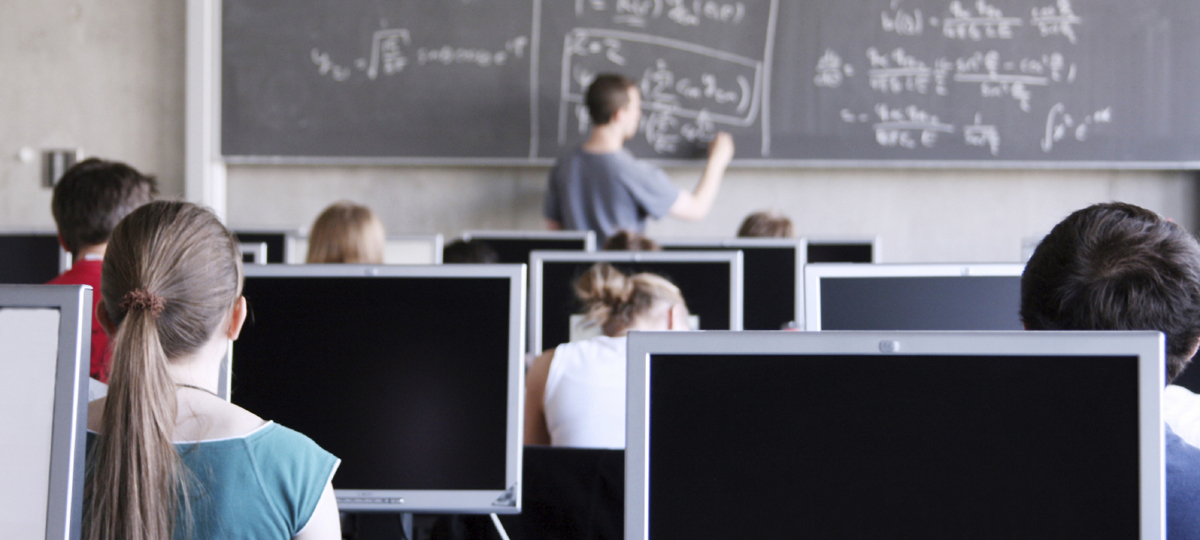 Students in a computer classroom, with math instructor writing on chalkboard