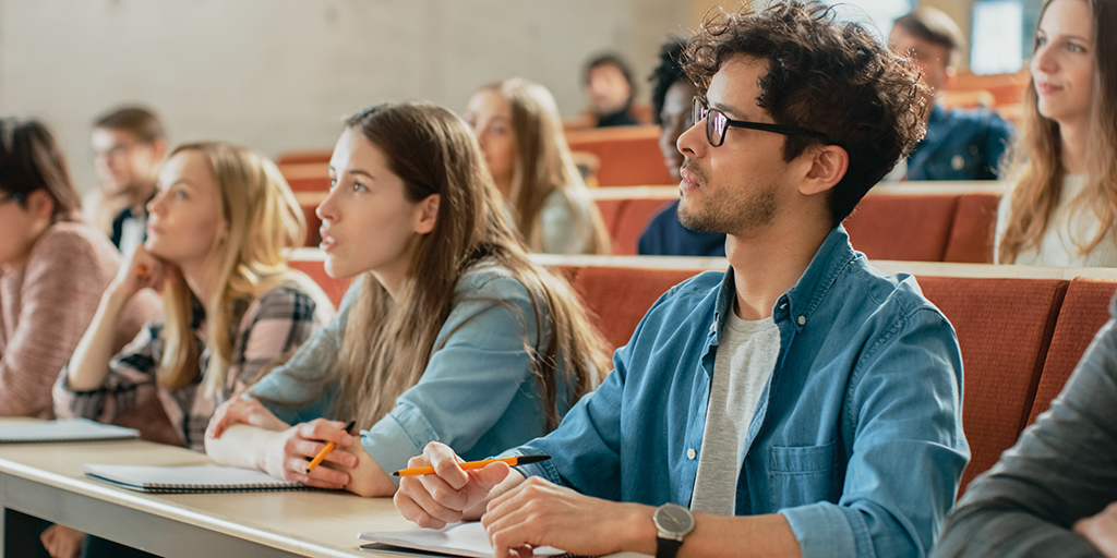 Students listen to an instructor in a classroom. The students, who appear college-aged, have notebooks and pencils.