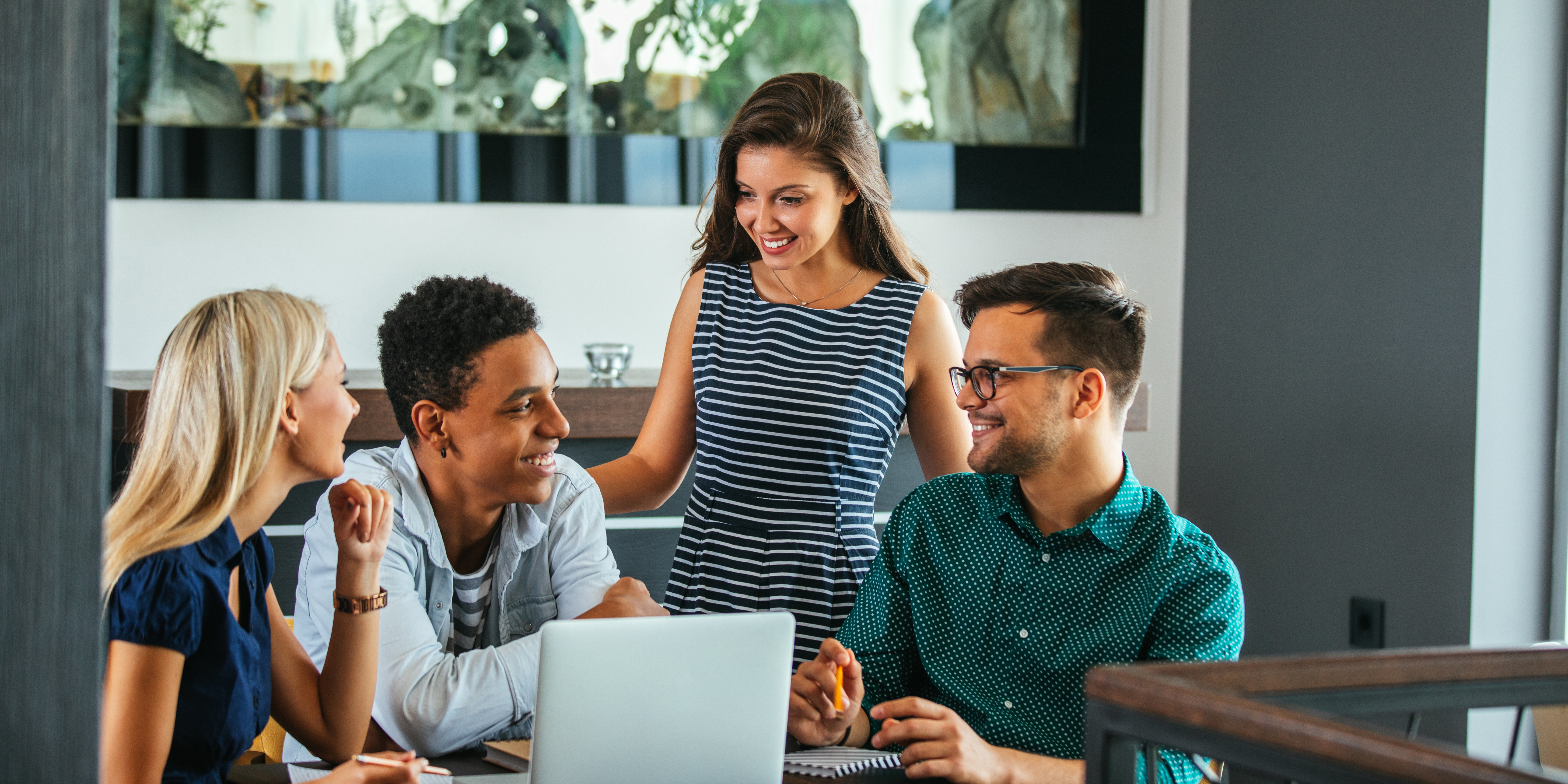 A group of three students sit around a laptop. A professor is behind them offering feedback