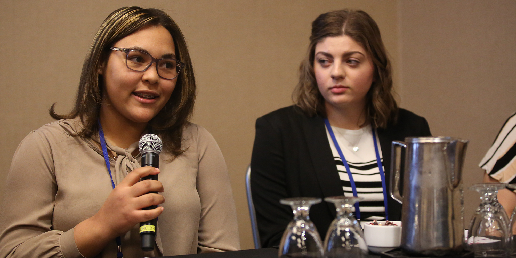 Two women sit behind a table. One holds a microphone