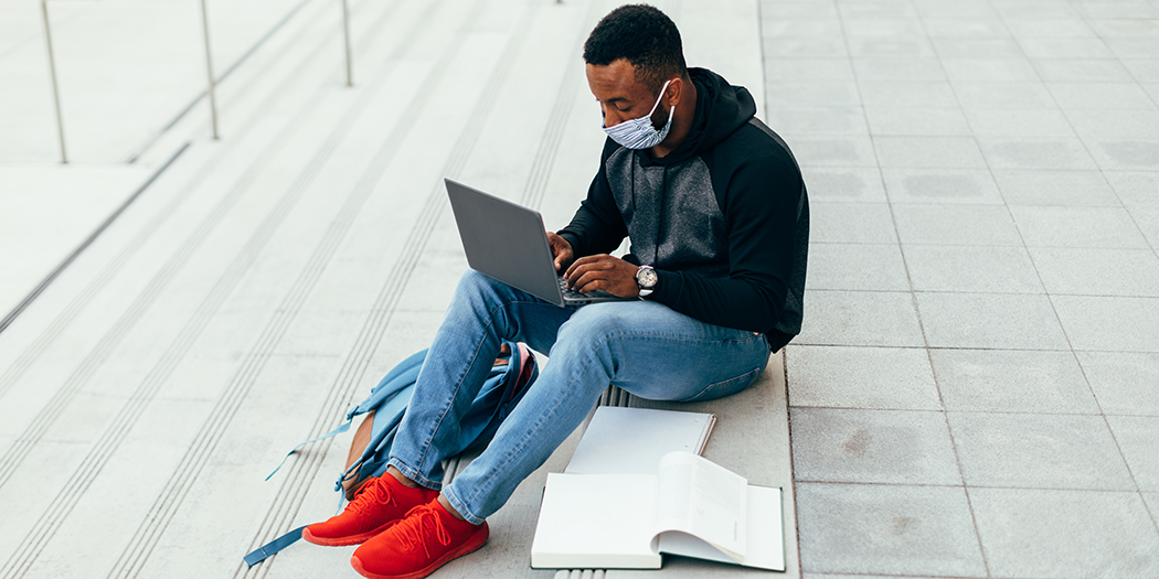 A college student wearing a mask sits with open books and a laptop