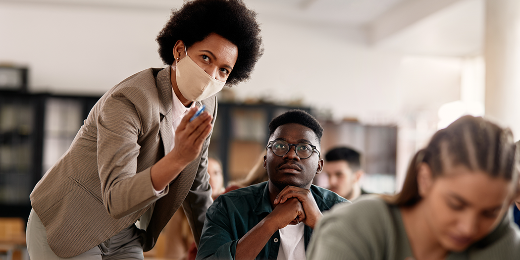 A professor wearing a mask helps a student in class