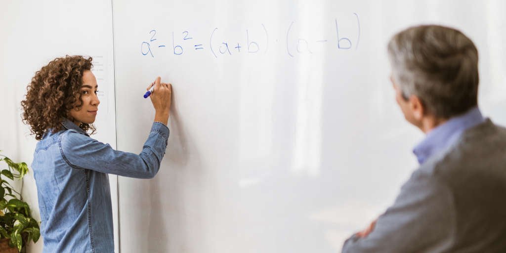 Student writing a formula on whiteboard while the teacher is observing.